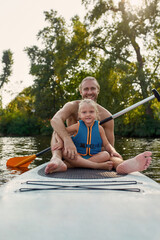 Portrait of father together with his little daughter smiling at camera, relaxing, sup surfing on a river on a summer day