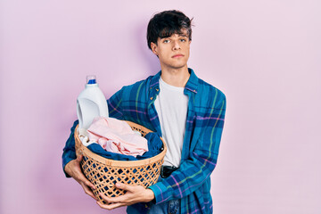 Poster - Handsome hipster young man holding laundry basket and detergent bottle relaxed with serious expression on face. simple and natural looking at the camera.