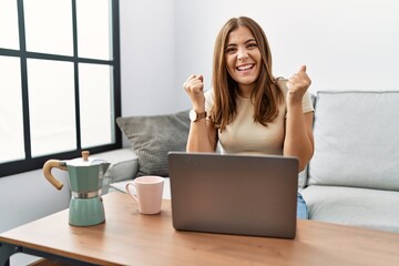 Canvas Print - Young brunette woman using laptop at home drinking a cup of coffee celebrating surprised and amazed for success with arms raised and open eyes. winner concept.