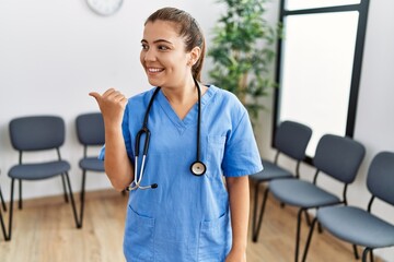 Sticker - Young brunette doctor woman at waiting room smiling with happy face looking and pointing to the side with thumb up.