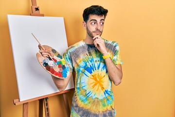 Young hispanic man standing drawing with palette by painter easel stand thinking worried about a question, concerned and nervous with hand on chin