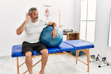 Sticker - Middle age hispanic man at pain recovery clinic holding pilates ball smiling with hand over ear listening an hearing to rumor or gossip. deafness concept.