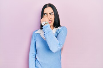 Poster - Beautiful woman with blue eyes standing over pink background smelling something stinky and disgusting, intolerable smell, holding breath with fingers on nose. bad smell