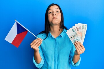 Poster - Young latin woman holding czech republic flag and koruna banknotes puffing cheeks with funny face. mouth inflated with air, catching air.