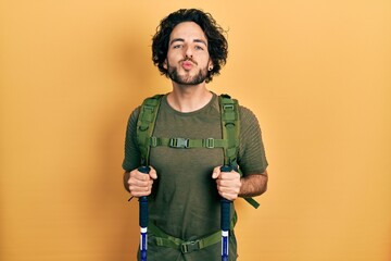 Poster - Handsome hispanic man wearing backpack holding trekking poles looking at the camera blowing a kiss being lovely and sexy. love expression.