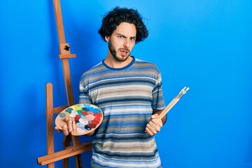 Poster - Handsome hispanic man standing drawing with palette by painter easel stand clueless and confused expression. doubt concept.