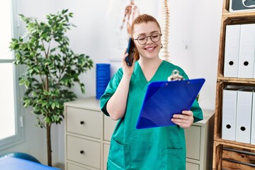 Wall Mural - Young redhead woman wearing phsiologist uniform talking on the smartphone at clinic