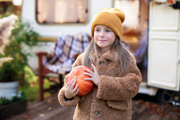 Happy Halloween. Smiling girl standing on the porch with a pumpkin in her hands. Trick-or-treat. Happy little girl playing near camping at RV home in fall garden. Child in cozy campsite fall backyard.