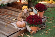 Happy Little Girl Sitting On Porch Of House With Chrysanthemum Potted And Pumpkins. Home Fall Decoration For Halloween Or Thanksgiving. Smilling Child In Autumn Garden With Yellow Pumpkins And Flowers