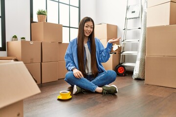 Sticker - Young chinese girl sitting on the floor at new home smiling cheerful presenting and pointing with palm of hand looking at the camera.