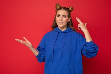 Wall Mural - Portrait of dissatisfied tired young beautiful winsome blonde woman with two horns with sincere emotions wearing stylish bright blue hoodie isolated on red background with copy space and showing gun