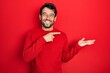 Handsome man with beard wearing casual red sweater amazed and smiling to the camera while presenting with hand and pointing with finger.