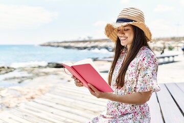 Poster - Young latin girl wearing summer hat reading book sitting on the bench at the beach.