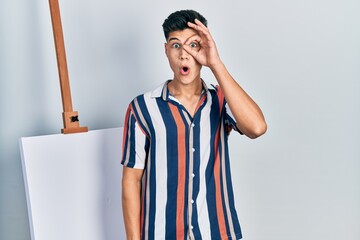 Canvas Print - Young hispanic man standing close to empty canvas doing ok gesture shocked with surprised face, eye looking through fingers. unbelieving expression.