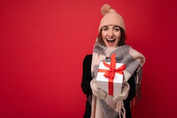 Fascinating happy joyful smiling adult brunette woman isolated over red background wall wearing winter scarf hat and mittens holding white gift box with red ribbon and looking at camera