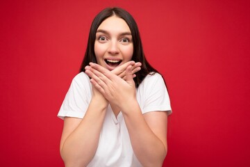 Wall Mural - Photo of positive young beautiful brunette female person with sincere emotions wearing casual white t-shirt for mockup isolated over red background with copy space and covering mouth