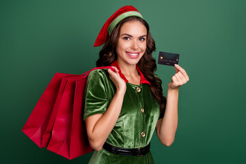 Poster - Portrait of beautiful trendy cheerful girl elf holding bank card carrying buyings isolated over green color background