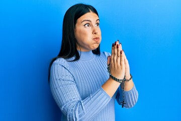 Poster - Beautiful brunette woman praying holding catholic rosary puffing cheeks with funny face. mouth inflated with air, catching air.