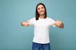 Portrait of positive happy smiling young beautiful brunette woman with sincere emotions wearing casual white t-shirt for mockup isolated over blue background with copy space and pointing at empty