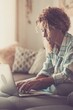 © simona - Beautiful young woman working on laptop sitting on sofa. Businesswoman working from home. Young woman in casuals sitting on couch. Woman using laptop at home