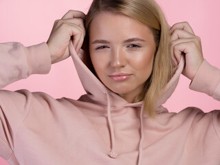 A cute young blonde in a pink hoodie puts a hood on her head.