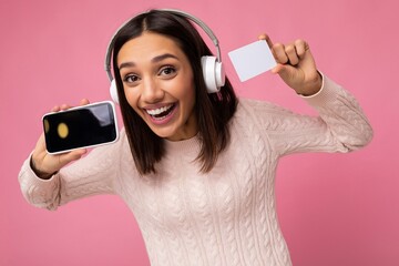 Wall Mural - Closeup portrait of beautiful overjoyed young brunette woman wearing pink casual sweater isolated over pink background wall wearing white bluetooth wireless headphones and listening to music and