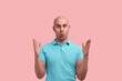 Impressed bald homosexual man with bristle shows big size with both hands, shapes huge object, has eyes wide open, pulls lips, gay friendly, wears blue polo shirt, stands over pink background.