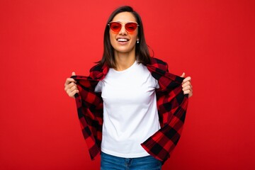 Portrait of positive cheerful smiling young brunette woman in casual white t-shirt for mockup, stylish red check shirt and trendy red sunglasses isolated on red background with copy space