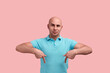 Confident serious bald unshaven homosexual man indicates with both index fingers down, demonstrates copy space for advertising content, gay friendly, wears blue polo shirt, stands on pink background.