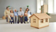 © Studio Romantic - Closeup small little wooden toy house on table, whole big family of mom, dad, grandma and grandpa sitting together on sofa in living-room in blurred background. Real estate and buying new home concept