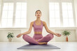 © Studio Romantic - Calm relaxed healthy young woman doing meditation exercise sitting in easy cross legged lotus asana on yoga mat on floor in modern white gym interior with bright light coming through large windows