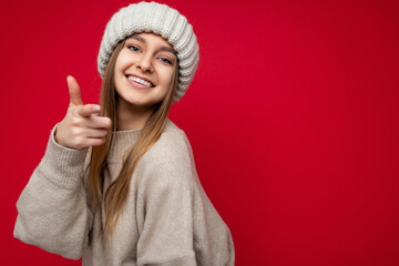 Wall Mural - Portrait of positive happy smiling young attractive dark blonde woman with sincere emotions wearing beige pullover and knitted hat isolated over red background with copy space and pointing at camera