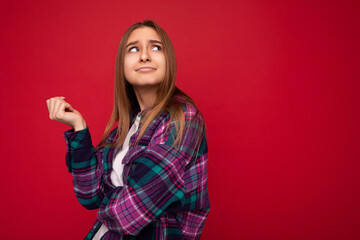 Photo of thoughtful young beautiful dark blonde woman with sincere emotions wearing hipster purple check shirt isolated over red background with copy space and thinking