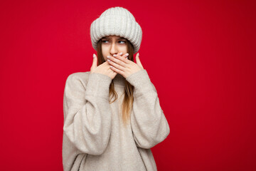 Wall Mural - Portrait of angry dissatisfied young nice pretty dark blonde female person with sincere emotions wearing beige jerseyr and beige hat isolated over red background with copy space and covers mouth with