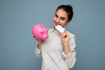 Portrait of happy positive smiling young beautiful brunette woman with sincere emotions wearing casual white shirt isolated on blue ackground with copy space, holds pink piggy box for money and credit