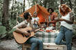 Group of young friends traveling in glamping in the forest having fun playing guitar and mbira kalimba roasting sausages sitting at dinner table near tent during summer vacation laughing hanging out