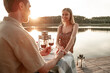 Adorable couple spend time together drinking wine on wooden pier: handsome man holds his girlfriend hand, making marriage proposal, wearing engagement ring, forest lake background