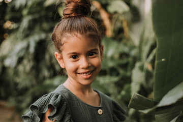 Wall Mural - The face of a little girl surrounded by tropical leaves. Closeup portrait of a beautiful swarthy baby with perfect skin and dark hair. Natural cosmetics, health, cleanliness, skin care, beauty concept