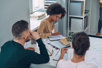 Three young modern people in smart casual wear communicating while working in the office together