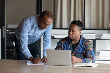 Serious African American adult mentor training young millennial intern at laptop, explaining. Boss explaining work task to new employee at workplace. Coworkers discussing project issues in office