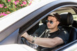 © Vladimir Razgulyaev - a teenager is driving. a modern young guy with sunglasses, fashionable with a beard, drives a white car. driving training or car travel