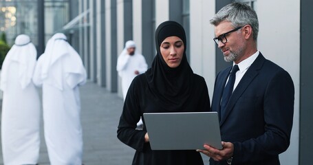 Wall Mural - Waist up of multi-ethnic professional managers working near the modern office. Caucasian male manger looking at the laptop screen and talking while muslim woman listening him attentively