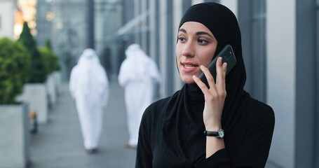 Wall Mural - Portrait view of the serious arabian businesswoman wearing head scarf standing at the street and chatting via smartphone with her business partners