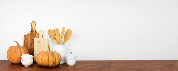 Wall Mural - Autumn theme kitchenware and utensils with rustic pumpkins on a wooden shelf or counter. Banner against a white wall background with copy space. Fall home kitchen cooking decor.