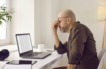 Tired Bald Man Has Eye Strain Because Of Constant Work On Laptop. Stressed Man Takes Off Glasses, Closes Irritated Eyes And Rubs Nose Bridge While Sitting At Desk With Notebook Computer In Home Office