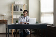 © fizkes - Happy young Indian doctor, therapist, general practitioner at work table with laptop in office, looking at camera, smiling, writing notes, medical records. Medic care concept. Professional portrait