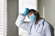 © fizkes - Exhausted young doctor feeling fatigue, stress due to work in covid hospital. Tired African mixed race practitioner in medical face mask, protective gloves wiping sweat from forehead with closed eyes.