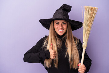 Wall Mural - Young Russian woman disguised as a witch holding a broom isolated on purple background smiling and raising thumb up