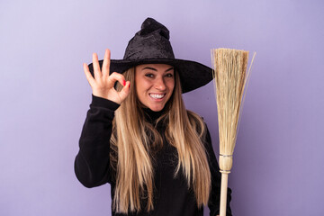 Wall Mural - Young Russian woman disguised as a witch holding a broom isolated on purple background cheerful and confident showing ok gesture.