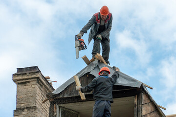 Repair of a wooden roof after a disaster against the background of a blue sky. An employee with a chainsaw in his hand and in special clothes is working on the roof. Violation of working conditions.